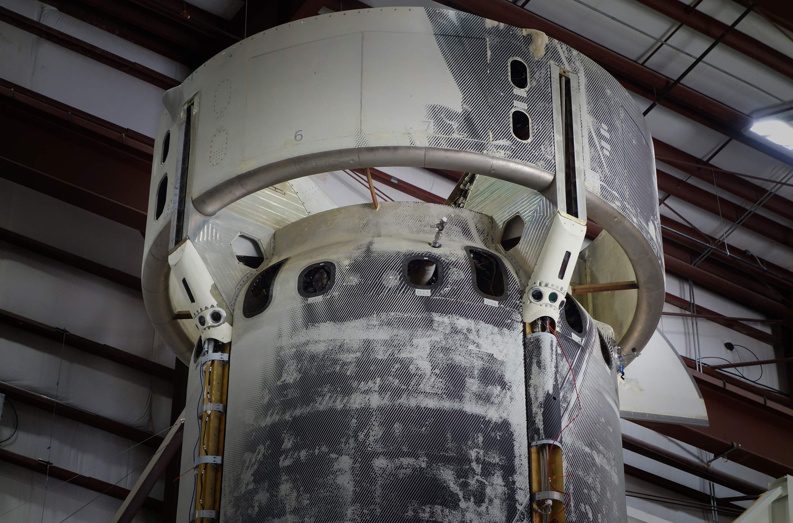 Industrial interior of a large white cylindrical spacecraft component under construction in a hangar, grayscale tones