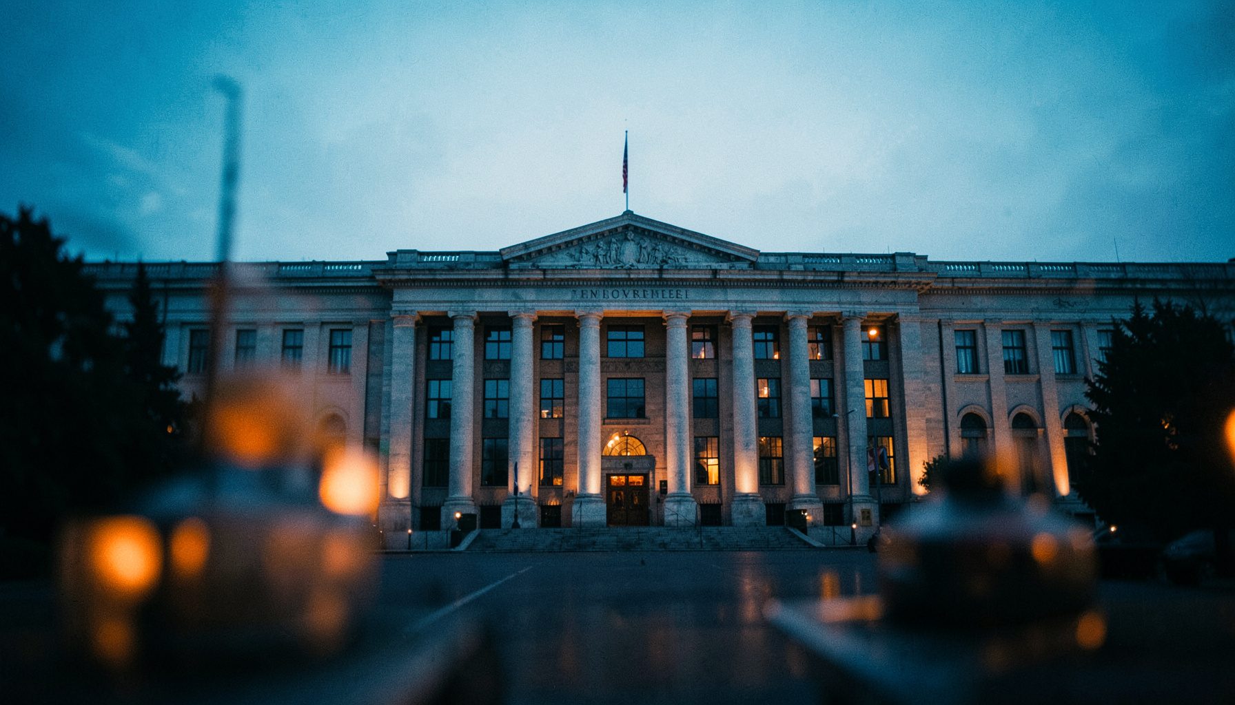 Wide cinematic frame of a government building facade at dusk, cool blue and amber tones, blurred foreground elements