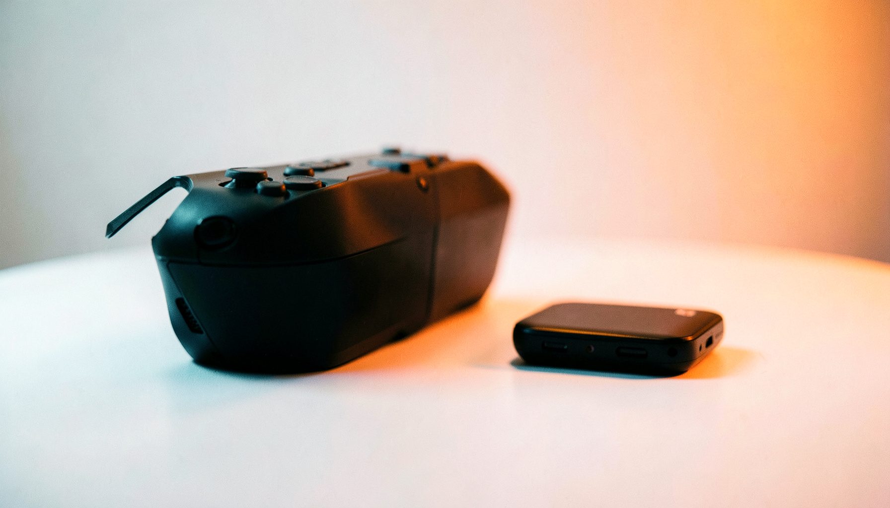 Two gaming consoles side by side on a minimalist white surface, one larger and one compact, soft gradient lighting